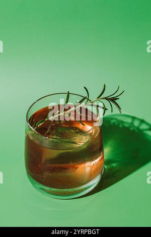 Foto eines dunkelbraunen Cocktails in einem Steinglas, garniert mit einem Zweig Rosmarin, vor grünem Hintergrund. Stockfoto