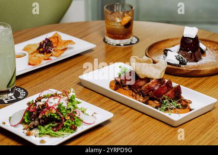 Ein vier-Gänge-Menü auf weißen Tellern und Craft-Cocktails, die am Rand eines Holztisches in einem modernen Café platziert werden. Stockfoto