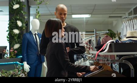 Modeexperte, die eine Vielzahl von trendigen Artikeln für den Kunden auswählen und ihm helfen, sich in der Umkleidekabine umzuziehen. Junger Mann, der Kleidung aus der neuen Kollektion anprobiert, Showroom. Kamera A.. Stockfoto