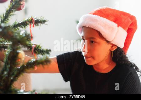 Kleine Brünette Latina Girl, die den Weihnachtsbaum sorgfältig dekoriert, während sie eine Weihnachtsmütze trägt. Stockfoto