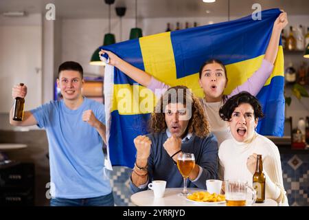 Eine Gruppe glücklicher Freunde, Sportfans, die ihre Lieblingsmannschaft mit der Flagge Schwedens anfeuern, während sie sich im Pub im Fernsehen ein Spiel anschauen Stockfoto