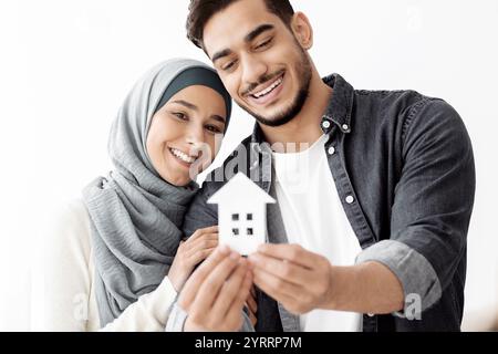 Junges, liebevolles muslimisches Paar, das Papierhaus hält und lächelt Stockfoto