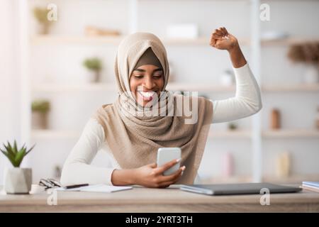 Überglücklich schwarze muslimische Frau in Hijab Blick auf Smartphone und feiert Erfolg Stockfoto