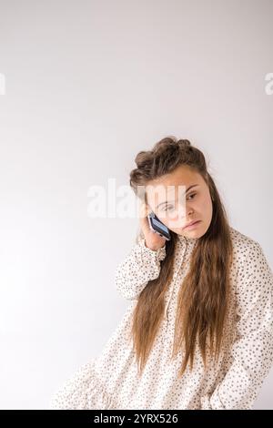 Eine junge Frau mit Down-Syndrom sitzt vor einem weißen Hintergrund und hält nachdenklich ein Handy an ihr Ohr. Sie trägt ein hübsches, florales d Stockfoto