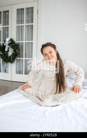 Ein junges Mädchen mit Down-Syndrom sitzt auf einem Bett und lächelt warm in die Kamera. Sie trägt ein hübsches, gemustertes Kleid. Das Bild vermittelt ein Gefühl der Freude Stockfoto