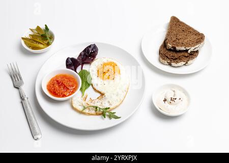 Teller mit Spiegelei, roter Soße und Basilikumblättern, weiße Porzellanschalen mit Käsesauce und eingelegten Gurken, Roggenbrotstücke auf der Untertasse Stockfoto