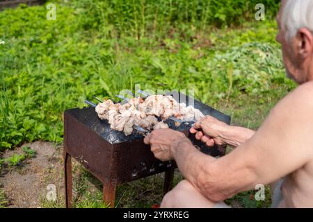 Ein älterer Mann grillt vorsichtig Fleischspieße über einer offenen Flamme in einem lebendigen und üppigen Hinterhof. Die Einstellung Stockfoto