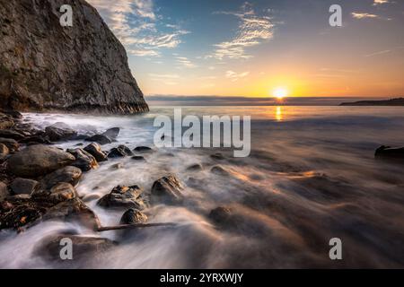 Wellen schlagen sanft gegen die felsige Küste, während die Sonne über Crystal Cove Beach untergeht und warmes Licht auf das ruhige Wasser und den Himmel wirft. Stockfoto