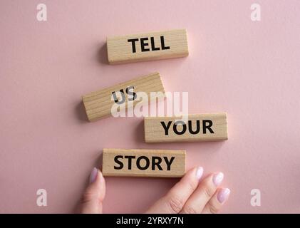 Erzählen Sie uns Ihr Story-Symbol. Konzeptworte erzählen uns Ihre Geschichte auf Holzblöcken. Schöner rosafarbener Hintergrund. Geschäftsmann Hand. Und sagen Sie uns, was Sie tun Stockfoto