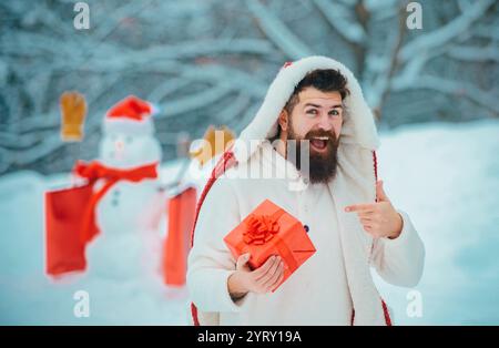 Weihnachten Winter Menschen Porträt. Mann mit rotem Geschenk, der mit Schneemann im Winterpark spielt. Schöner Weihnachtsmann. Schneemann und lustiger bärtiger Mann im sn Stockfoto