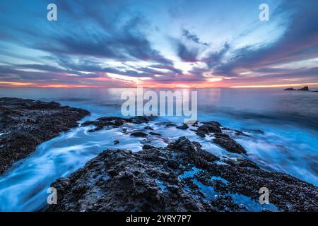 Leuchtende Farben erfüllen den Himmel, während die Sonne über dem ruhigen Wasser und der felsigen Küste von Crescent Bay Beach in Südkalifornien untergeht. Stockfoto