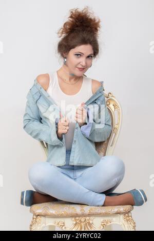 Eine schöne Frau in einem Denim-Anzug posiert auf einem Stuhl auf grauem Hintergrund. Stockfoto