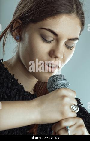 Junge Frau, die leidenschaftlich in ein Mikrofon singt, mit geschlossenen Augen, voll eingetaucht in die Emotionen ihres Auftritts. Das Wesen der Musik und einfangen Stockfoto