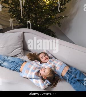 Zwei lächelnde Schwestern in passenden, gemütlichen Pullover entspannen sich auf einer hellgrauen Couch neben einem wunderschön verzierten Weihnachtsbaum. Stockfoto