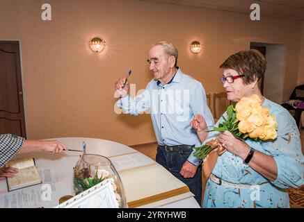 Ältere russische Paare in ihren 70ern feiern ihren 50. Hochzeitstag, indem sie Dokumente in einem Standesamt unterschreiben. Die Frau hält einen Strauß davon Stockfoto