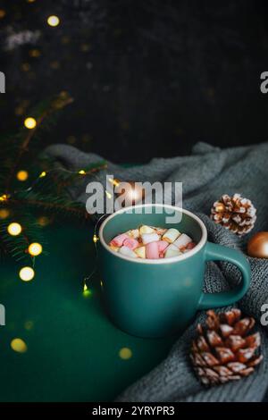 Eine Tasse heiße Schokolade mit Marshmallows, umgeben von Tannenzapfen, Feenlichtern und Weihnachtsdekor auf einem dunklen, gemütlichen Hintergrund, schaffen ein warmes und einladendes Ambiente Stockfoto