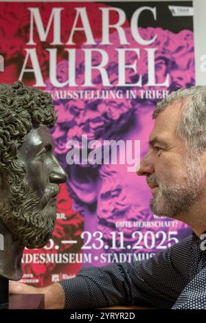 Trier, Deutschland. Dezember 2024. Marcus Reuter, Direktor des Rheinischen Landesmuseums in Trier, blickt in die Kamera vor einem Plakat zur Landesausstellung neben einer Nachbildung des Kopfes des römischen Kaisers Marcus Aurelius. Die Ausstellung im Rheinischen Landesmuseum und im Stadtmuseum Trier findet vom 15. Juni bis 23. November 2025 statt. Quelle: Harald Tittel/dpa/Alamy Live News Stockfoto