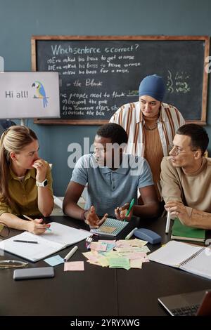 Vertikale Aufnahme des jungen Schwarzen Mannes, der mit einer Lehrerin spricht, die in Gruppendiskussionen am Schreibtisch sitzt, während er neue Wörter in der Sprechstunde in der Sprachschule für Erwachsene Lernende übt Stockfoto