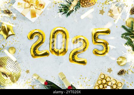 Neujahr 2025, kreative Idee. Goldene Ballons 2025 liegen auf einem grauen Betontisch mit Kaminen, Süßigkeiten, einer goldenen Discokugel, Champagner und einem Weihnachtsfest Stockfoto