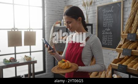 Eine junge Frau in einer Bäckerei, die eine rote Schürze trägt und ein Gebäck hält, während sie ihr Handy in einer gemütlichen Einrichtung ansieht Stockfoto