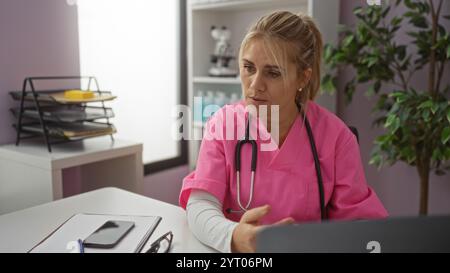 Frau in rosa Schrubben mit Stethoskop sitzt im Krankenhausbüro und unterhält sich mit dem Patienten drinnen neben dem Schreibtisch mit Akten und Smartphone Stockfoto