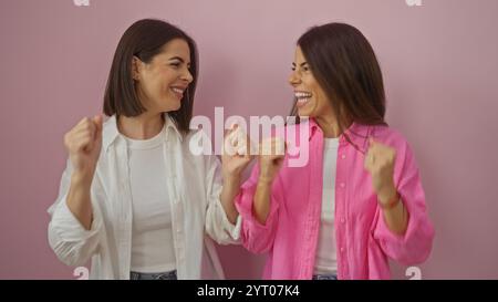 Zwei lächelnde Frauen in legeren Outfits feiern begeistert vor rosa Hintergrund und symbolisieren Freundschaft und Freude. Stockfoto
