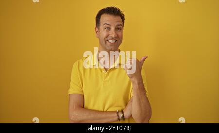 Hispanischer Mann mittleren Alters, der selbstbewusst in einem gelben Hemd lächelt und eine Geste nach oben gibt, isoliert vor einer leuchtend gelben Hintergrundwand. Stockfoto