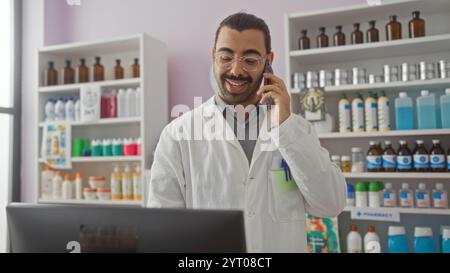Junger hispanischer Mann mit Schnurrbart und Brille, der in der Apotheke arbeitet, während er am Telefon neben den Arzneiregalen spricht Stockfoto