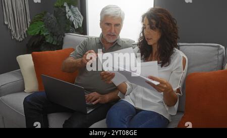 Ehepaar mittleren Alters diskutieren über Papierkram im Wohnzimmer, Mann mit Laptop, Frau mit Dokumenten, Schaffung einer fokussierten Arbeitsumgebung im Innenbereich. Stockfoto