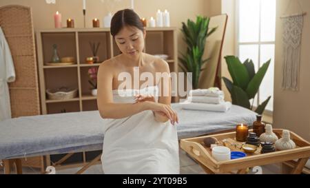 Junge Frau in einem Spa, die in einem ruhigen Schönheitssalon Creme auf den Arm aufträgt, mit Kerzen und Hautpflegeprodukten auf einem Tablett. Stockfoto