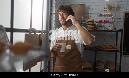 Hübscher junger Mann, der am Telefon spricht, während er eine Tasse in einer gemütlichen Bäckerei mit verschiedenen Gebäckstücken hält, eine Schürze trägt und fröhlich Indoo aussieht Stockfoto