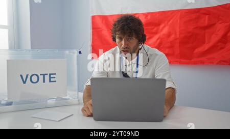 Junger Mann, der in einem Wahlzimmer mit einer polnischen Flagge im Hintergrund abstimmt, auf einen Laptop blickt und konzentriert erscheint. Stockfoto