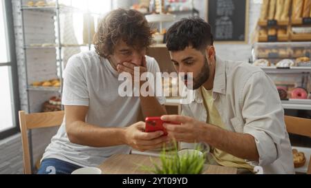 Zwei Männer sitzen an einem Tisch in einer Bäckerei und blicken auf einen roten Smartphone-Bildschirm mit Ausdrücken der Überraschung und Neugier Stockfoto