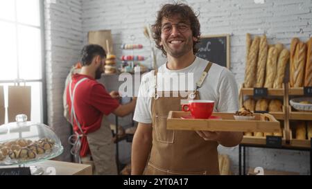 Zwei lächelnde Bäcker in Schürzen arbeiten drinnen in einer Bäckerei zusammen, wobei einer Kaffee und einen Cupcake serviert, während der andere Backwaren anlegt Stockfoto