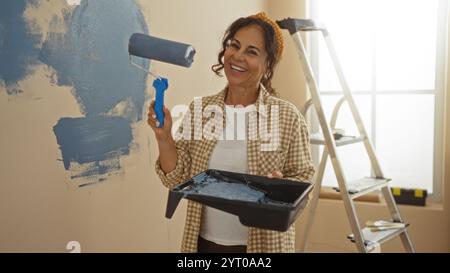 Frau malte die Wand in einem neuen Wohnraum mit Roller und Leiter, lächelnd und hielt Farbtablett, trug kariertes Hemd, helles Zimmer mit Sonnenlicht Stockfoto
