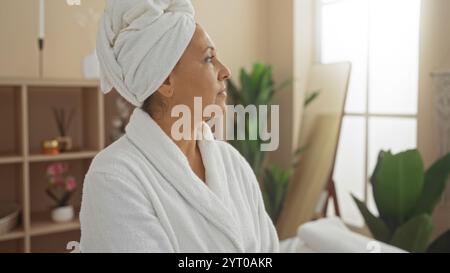 Eine wunderschöne hispanische Frau mittleren Alters genießt einen entspannenden Moment in einer friedlichen Spa-Umgebung, die in ein weißes Handtuch und einen Bademantel gehüllt ist und von Kalk umgeben ist Stockfoto