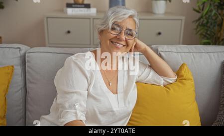 Grauhaarige Frau lächelt in einem gemütlichen Wohnzimmer mit gelben Kissen und zeigt eine schöne, reife Person mittleren Alters in einer Inneneinrichtung. Stockfoto