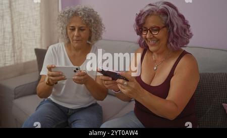 Zwei ältere Frauen mit grauen Frisuren spielen fröhlich Videospiele auf Smartphones, während sie zusammen auf einer Couch in einem gemütlichen Wohnzimmer sitzen. Stockfoto
