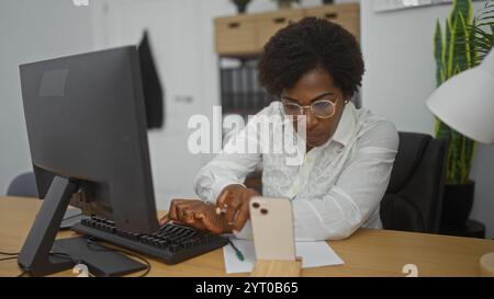 Frau, die in einem Büro arbeitet, auf ein Telefon schaut, während sie einen Computer benutzt, afroamerikaner, lockiges Haar, mittleres Alter, Profi, Brille, fokussieren auf ta Stockfoto