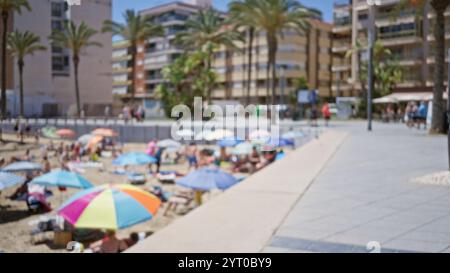 Verschwommenes Bild von Menschen, die an einem sonnigen Tag am Strand mit bunten Sonnenschirmen, Palmen und Gebäuden im Hintergrund entspannen Stockfoto
