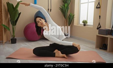 Frau dehnt sich drinnen auf einer Yogamatte in einem Fitnessstudio und demonstriert eine seitliche Dehnübung in einem ruhigen und gut beleuchteten Raum mit Pflanzen und Fitnessgeräten Stockfoto