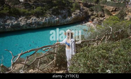Junge Frau, die den malerischen Blick auf cala del moro auf mallorca, spanien, genießt, in der Nähe von klarem türkisfarbenem Wasser und natürlichen Klippen unter dem sonnigen Himmel. Stockfoto