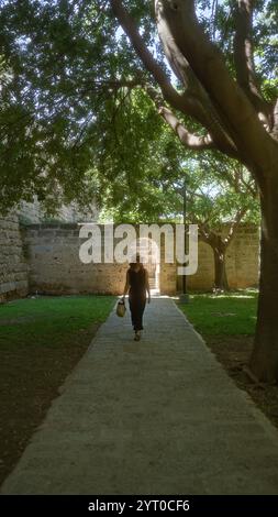 Frau, die durch einen schattigen historischen Innenhof in palma mallorca, spanien, auf einem von Steinmauern und üppigen grünen Bäumen gesäumten Weg geht und einen Strohbeutel trägt Stockfoto