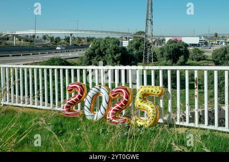 Einige Folienballons in verschiedenen Farben bilden die Zahl 2025, als das neue Jahr, gestützt auf die Gitter eines weißen Geländes in einem Industriepark, auf einem Su Stockfoto