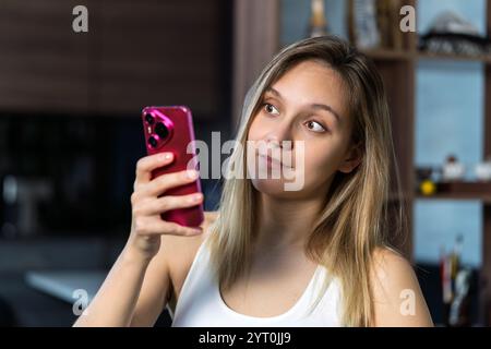 Ein Mädchen, das im Haus auf dem Smartphone schreibt und sie ist schockiert. Die blonde Frau benutzt ihr pinkfarbenes Smartphone. Schockierte schlechte Nachrichten Stockfoto