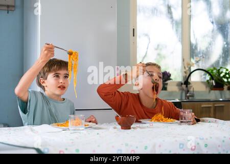 Zwei junge Brüder genießen eine Mahlzeit aus Spaghetti mit Tomatensauce, die verspielte und fröhliche Ausdrücke beim Essen in ihrer Küche zeigen Stockfoto