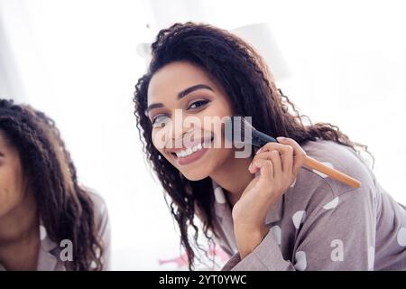 Junge Frau im Schlafanzug lächelt beim Auftragen von Make-up zu Hause bei natürlichem Licht Stockfoto
