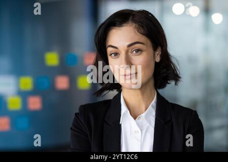 Selbstbewusste Geschäftsfrau mit kurzen schwarzen Haaren in professioneller Kleidung posiert in moderner Büroumgebung. Sie vermittelt Professionalität, Selbstvertrauen und Ehrgeiz Stockfoto