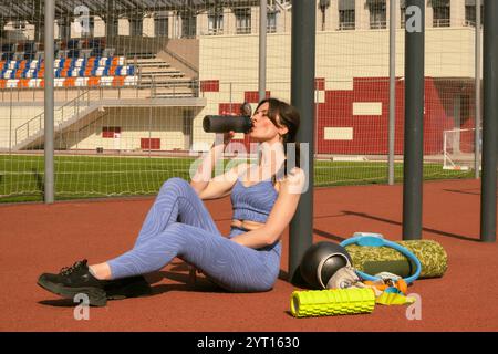 Die Frau in blauer Sportausrüstung sitzt mit einer Wasserflasche, umgeben von Fitnessgeräten, und reflektiert ihre Trainingseinheit. Dieses Foto hebt hervor Stockfoto