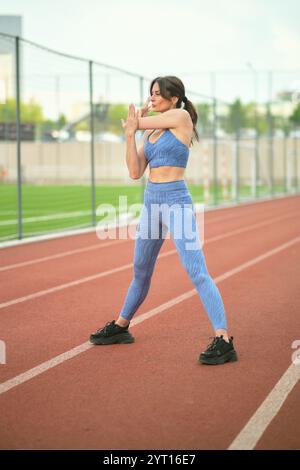 Eine Frau in blauer Sportbekleidung streckt ihren Arm auf einer Rennstrecke und bereitet sich auf ein Workout vor. Dieses Bild unterstreicht die Bedeutung von Aufwärmübungen für Stockfoto